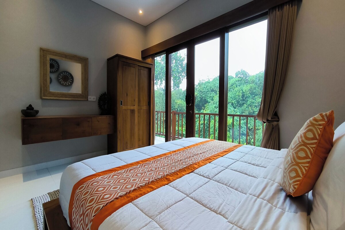 A bedroom with a large bed, a wooden wardrobe, a framed picture on the wall, and a balcony with a view of greenery.