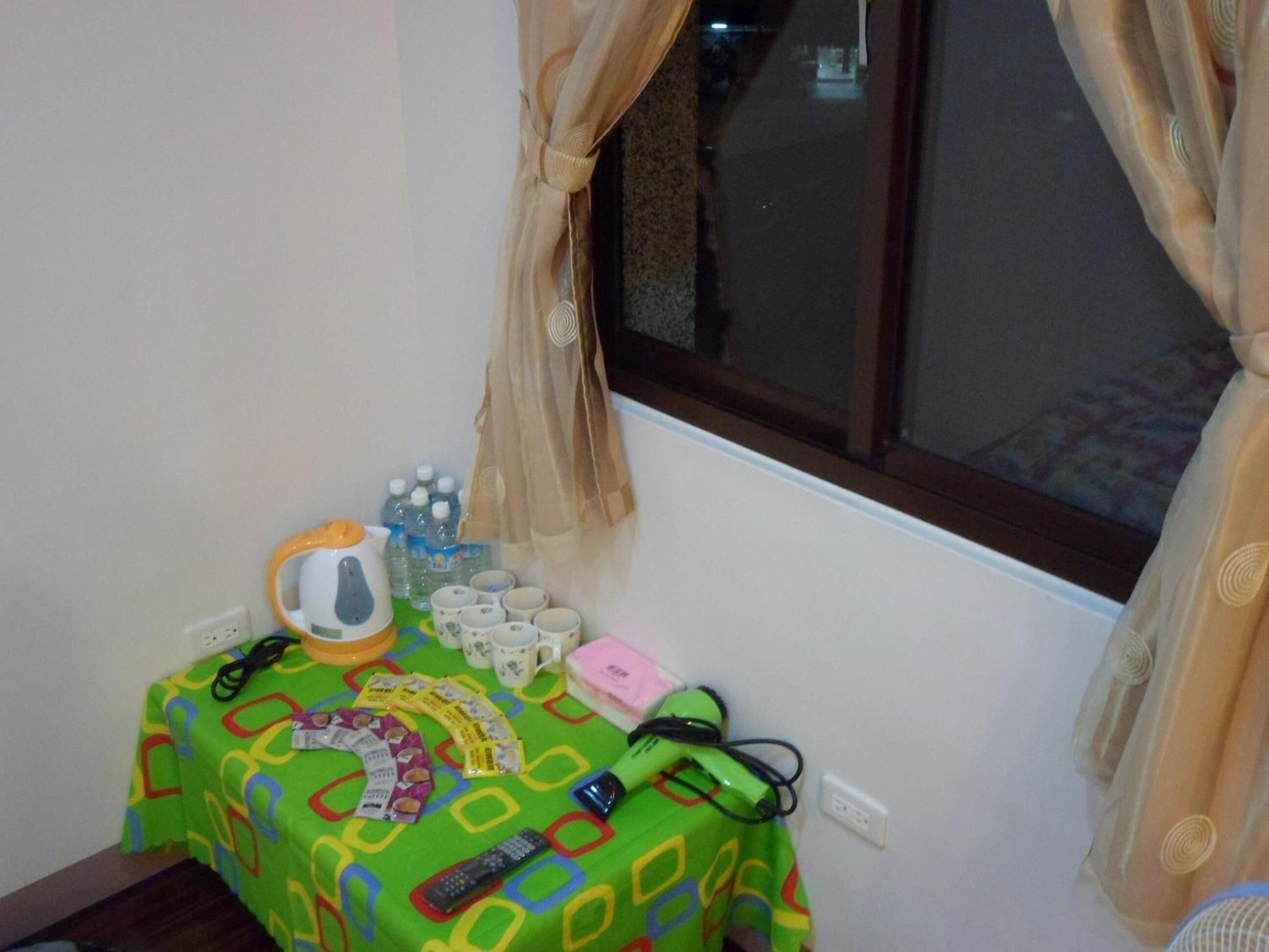 Una mesa pequeña con mantel colorido, una tetera, tazas, un control remoto y algunos bocadillos.