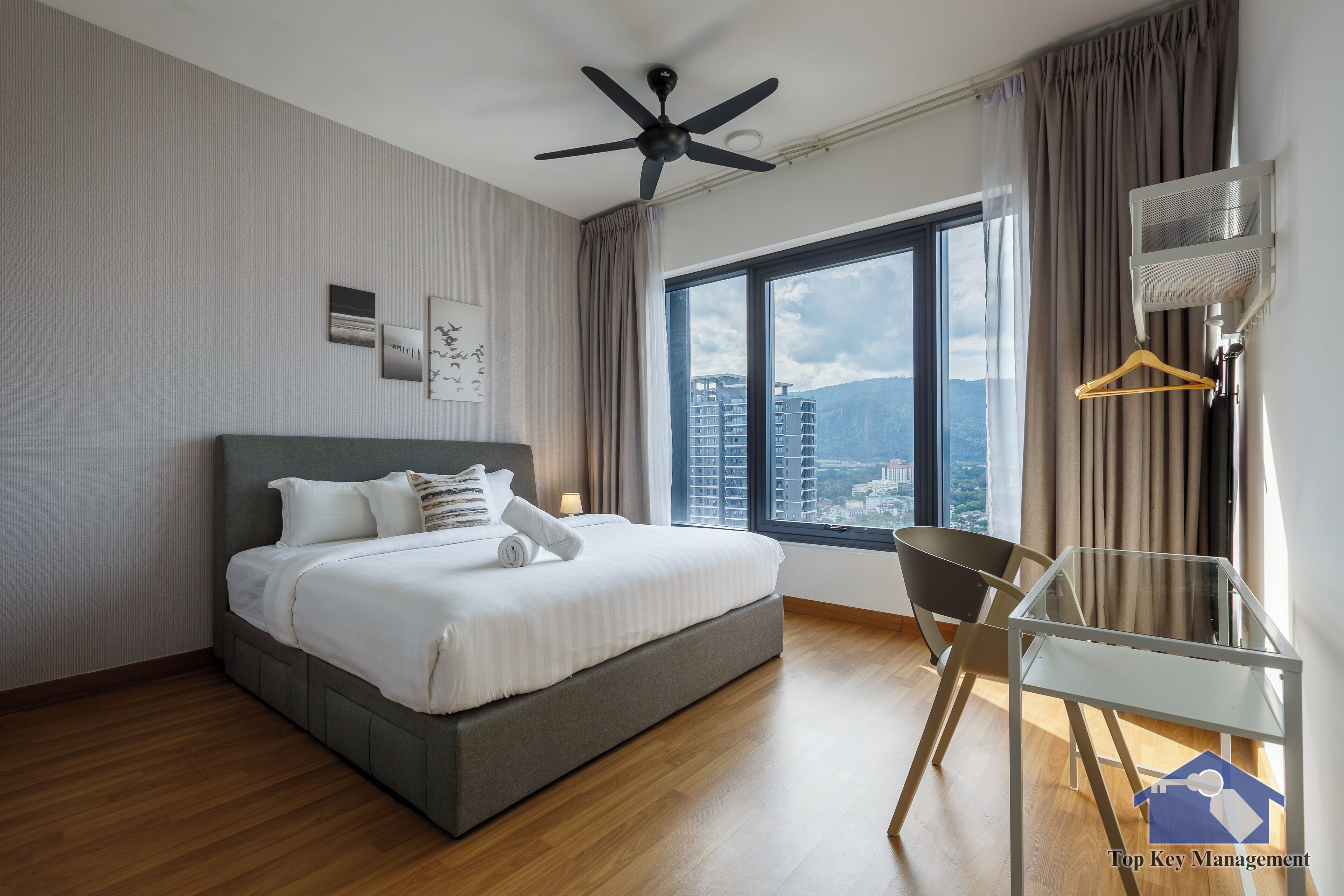 A modern hotel room with a large bed, a ceiling fan, a glass table, and a view of the city.