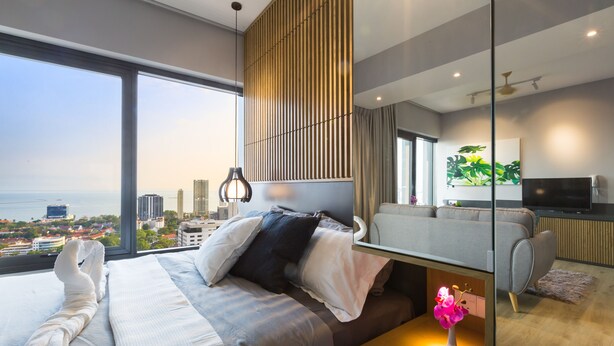 A modern hotel room with a large window, a bed with white linens, a grey sofa, a flat-screen TV, and a small table with a vase. - Tropicana 218 Macalister (George Town)