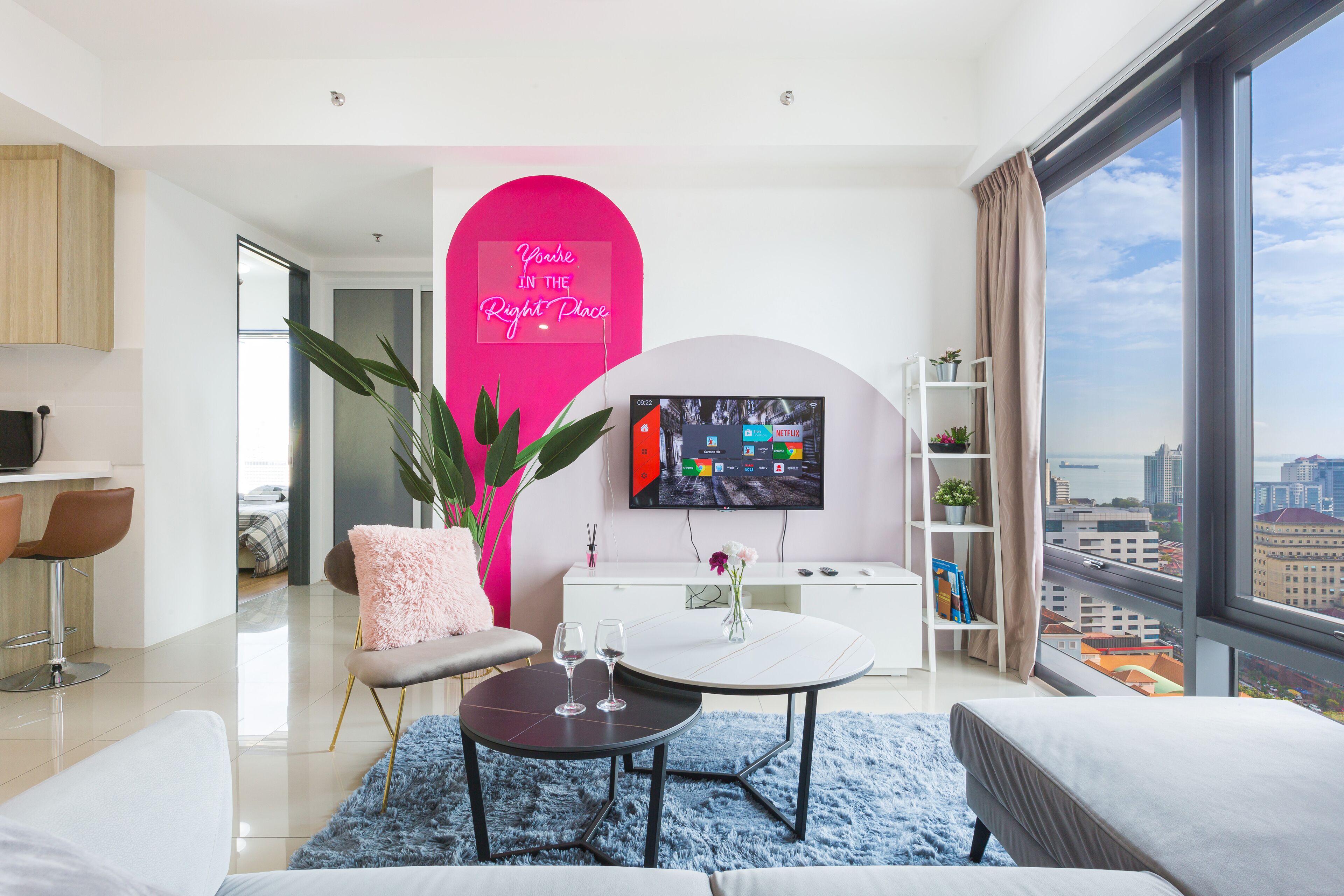 A modern living room with a large TV, a pink accent wall, and a city view.