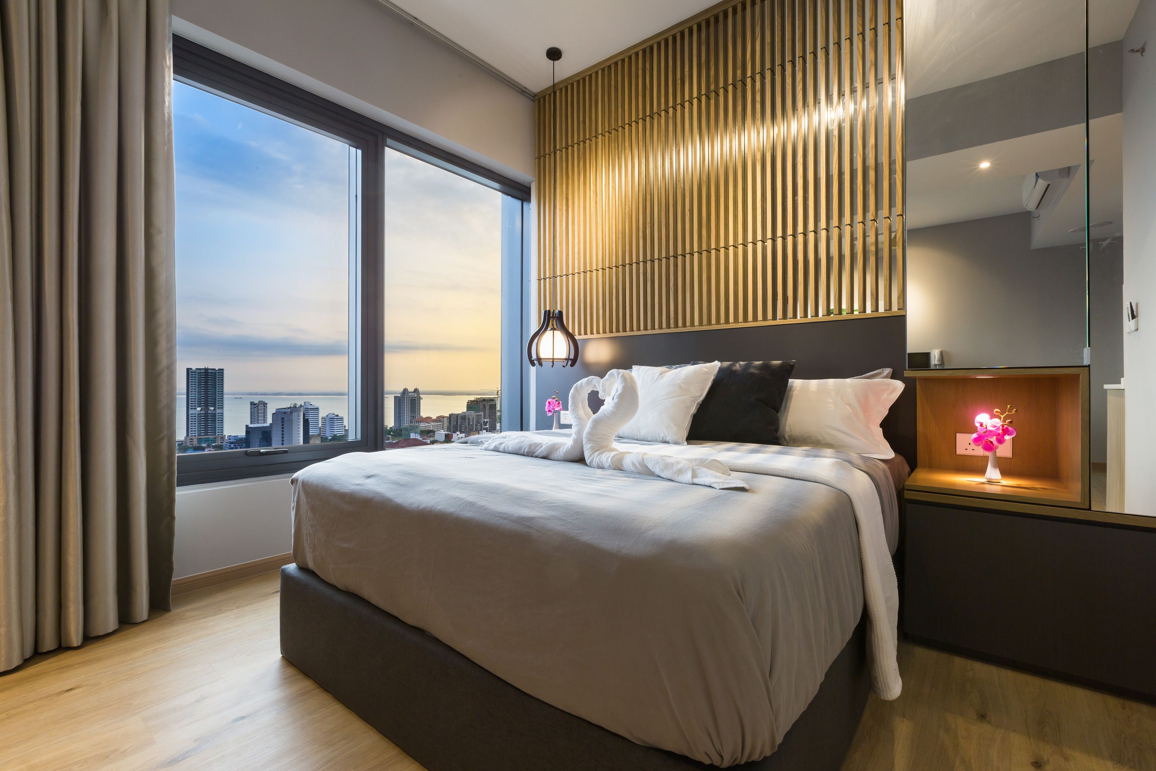 A modern hotel room with a large bed, a city view, a wooden slat headboard, and a bedside table with a lamp.