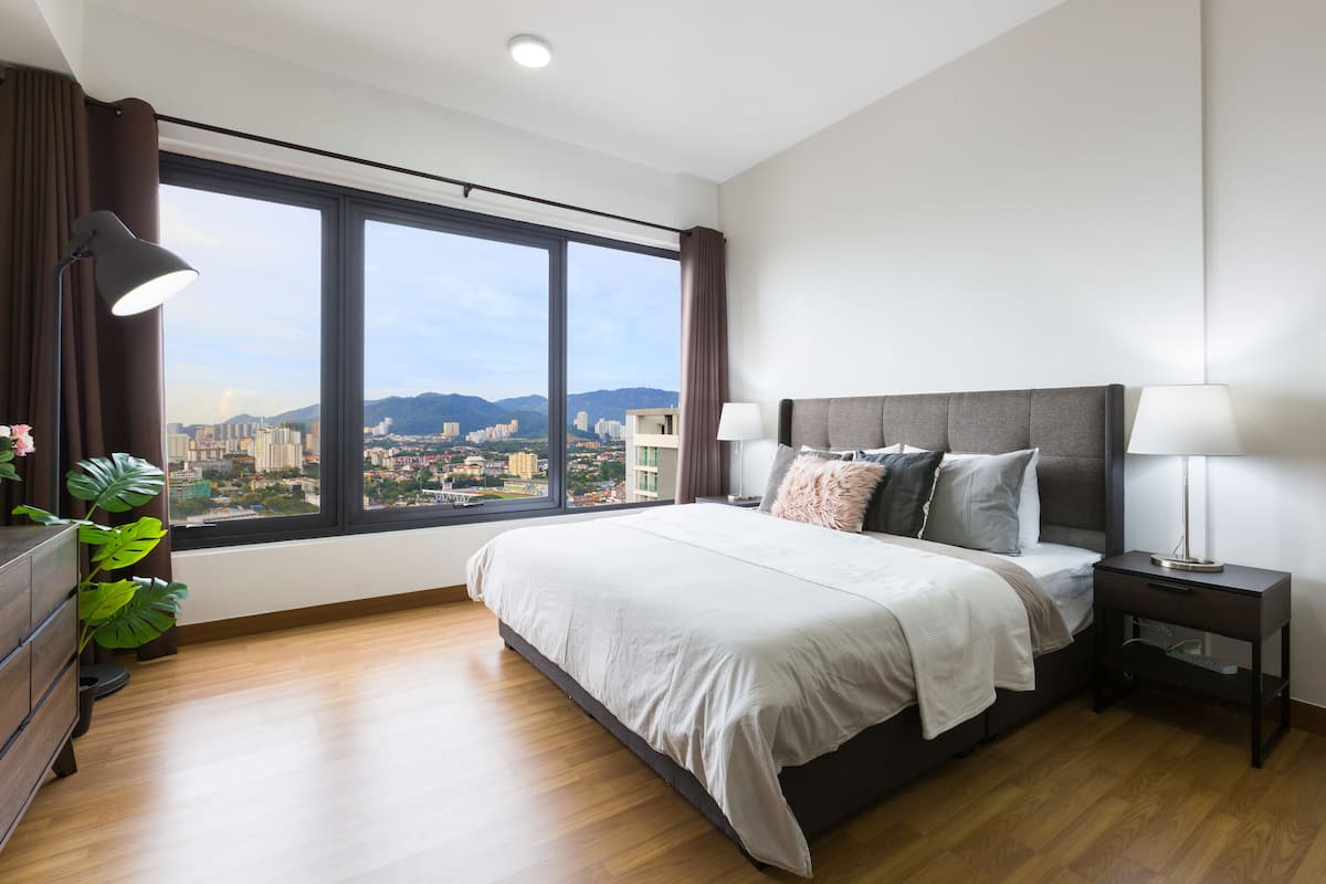 A modern hotel room with a large bed, a nightstand, a dresser, and a view of a cityscape through a large window.