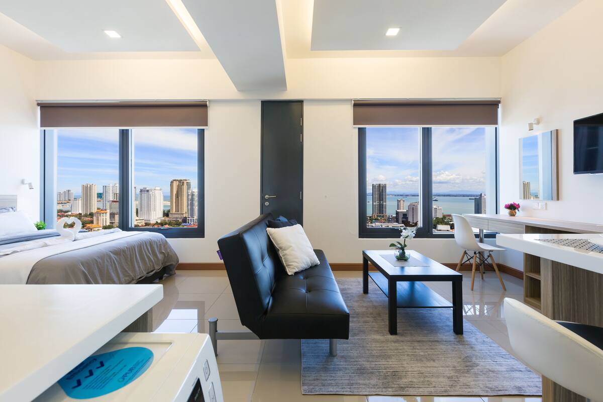 A modern hotel room with a bed, a black leather sofa, a desk, and large windows offering a city view.