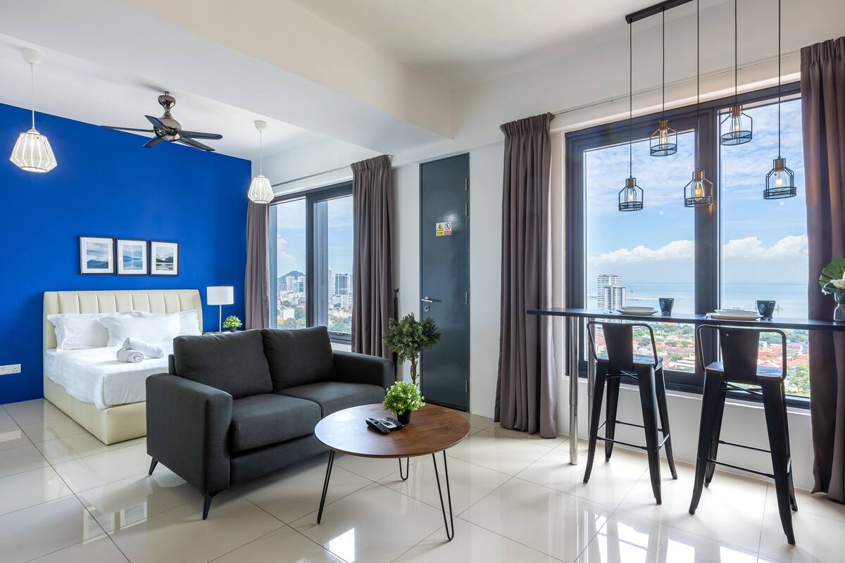 A modern hotel room with a bed, a sofa, a coffee table with a plant, and a dining area with bar stools.