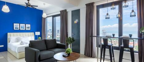 A modern hotel room with a bed, a sofa, a coffee table with a plant, and a dining area with bar stools.