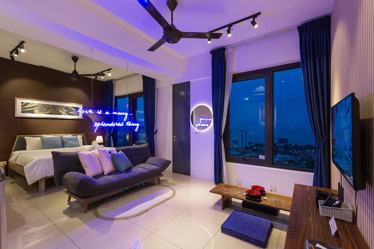 A modern living room with a sofa, a coffee table, and a TV. There is a bed with a headboard and a window with curtains.