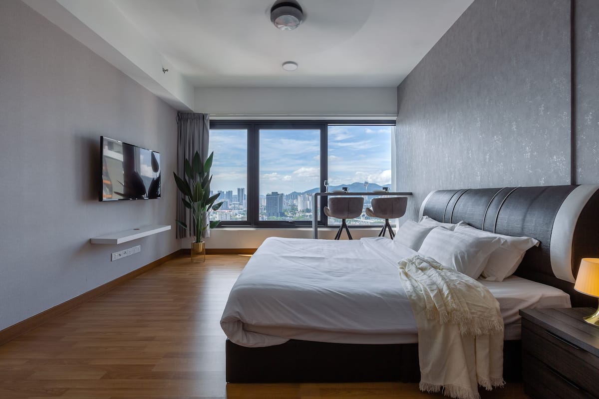 A modern hotel room with a large bed, a TV mounted on the wall, a city view through large windows, and a small seating area with a chair and table.