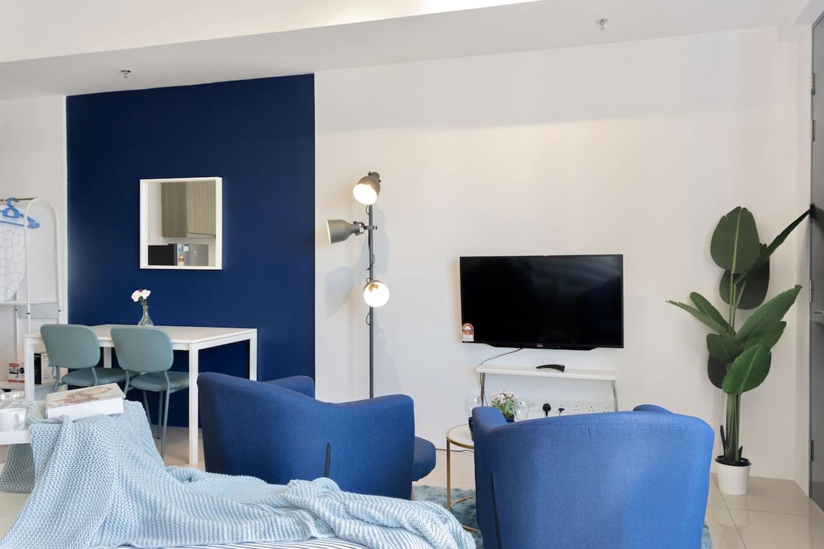 A modern living room with a blue accent wall, a flat-screen TV, a desk with chairs, and a large potted plant.