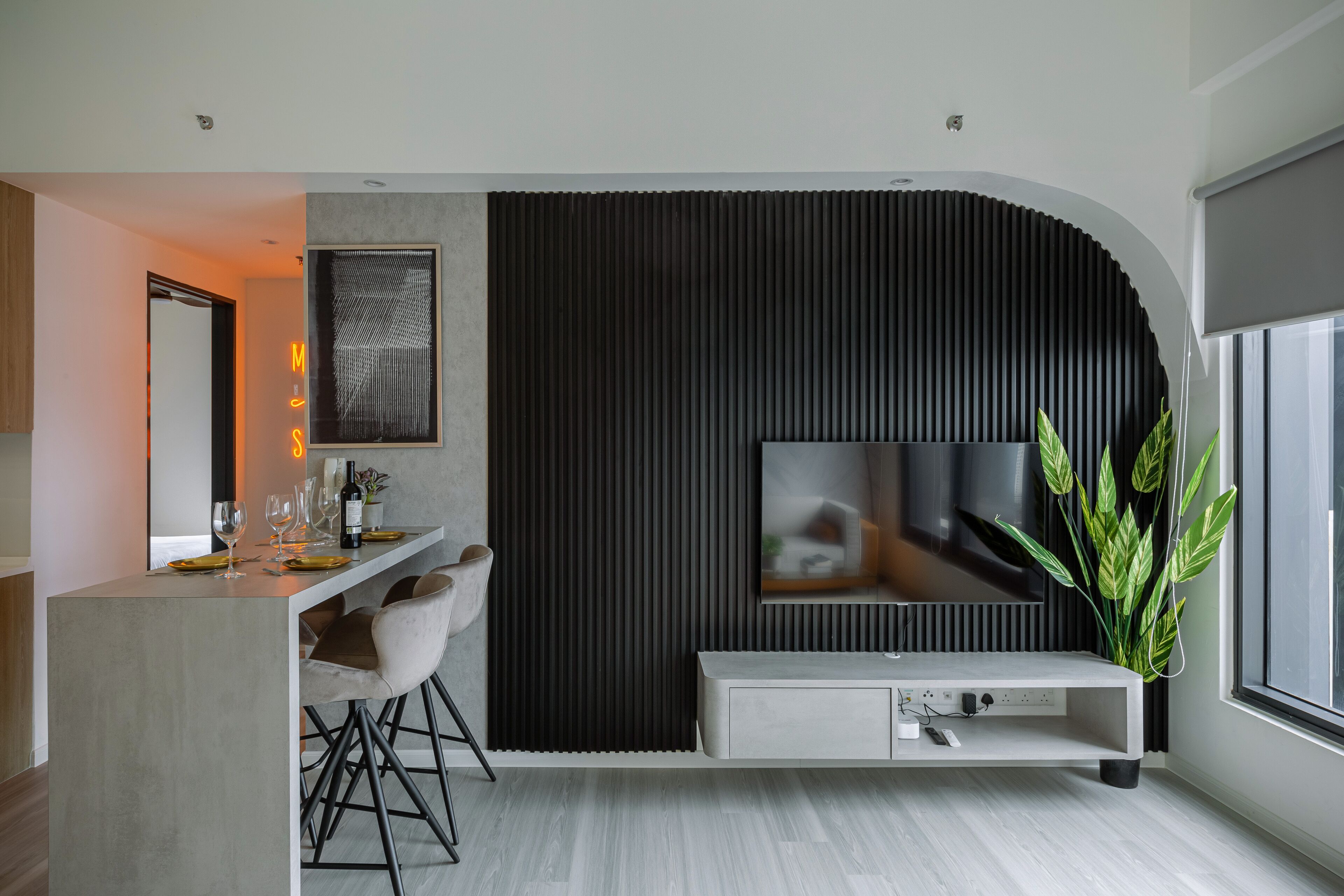 A modern living room with a TV mounted on a black wall, a bar area with stools, and a large plant.