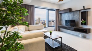 A modern living room with a sofa, coffee table, TV, and large windows offering a city view. - Tropicana 218 Macalister (George Town)