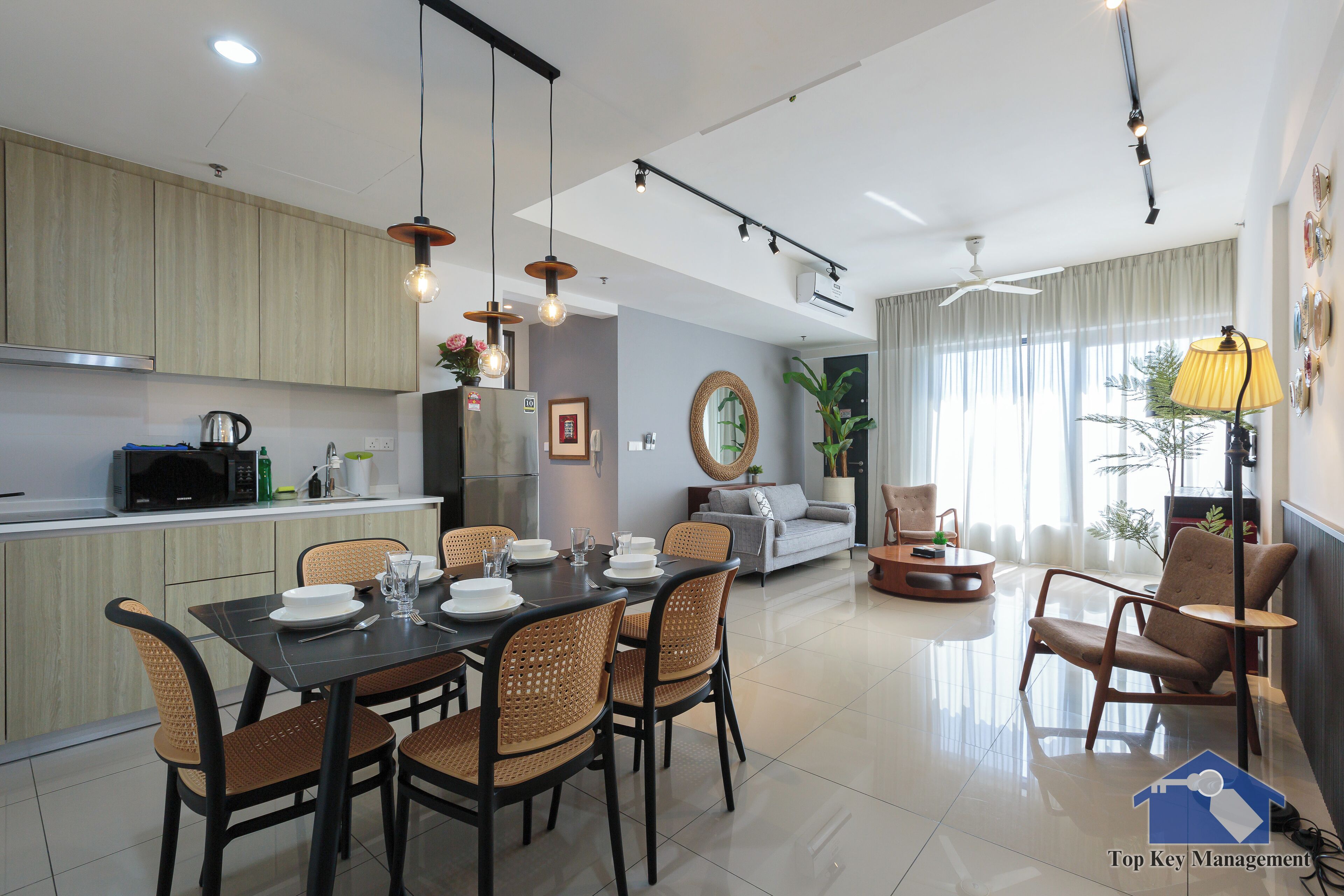 A modern kitchen with wooden cabinets, a dining area with a black table and wicker chairs, and a living room with a sofa and armchairs.