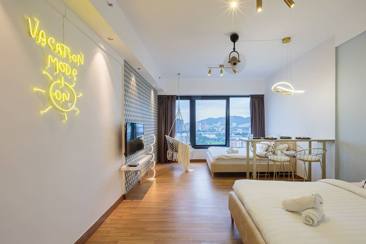 A modern hotel room with a large bed, a desk, and a TV. There's a neon sign on the wall and a view of the city through the window.