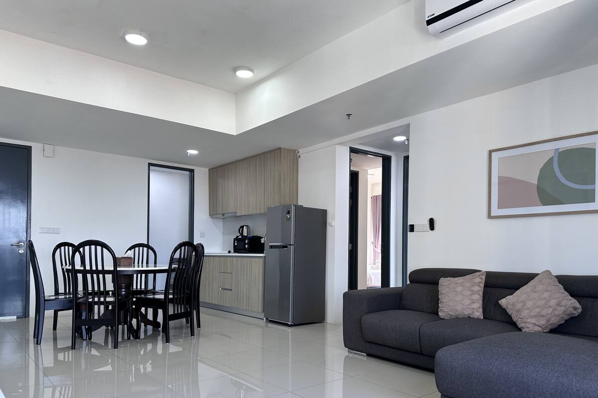 A modern living room with a grey sofa, a dining table with chairs, and a kitchen area with a refrigerator and cabinets.