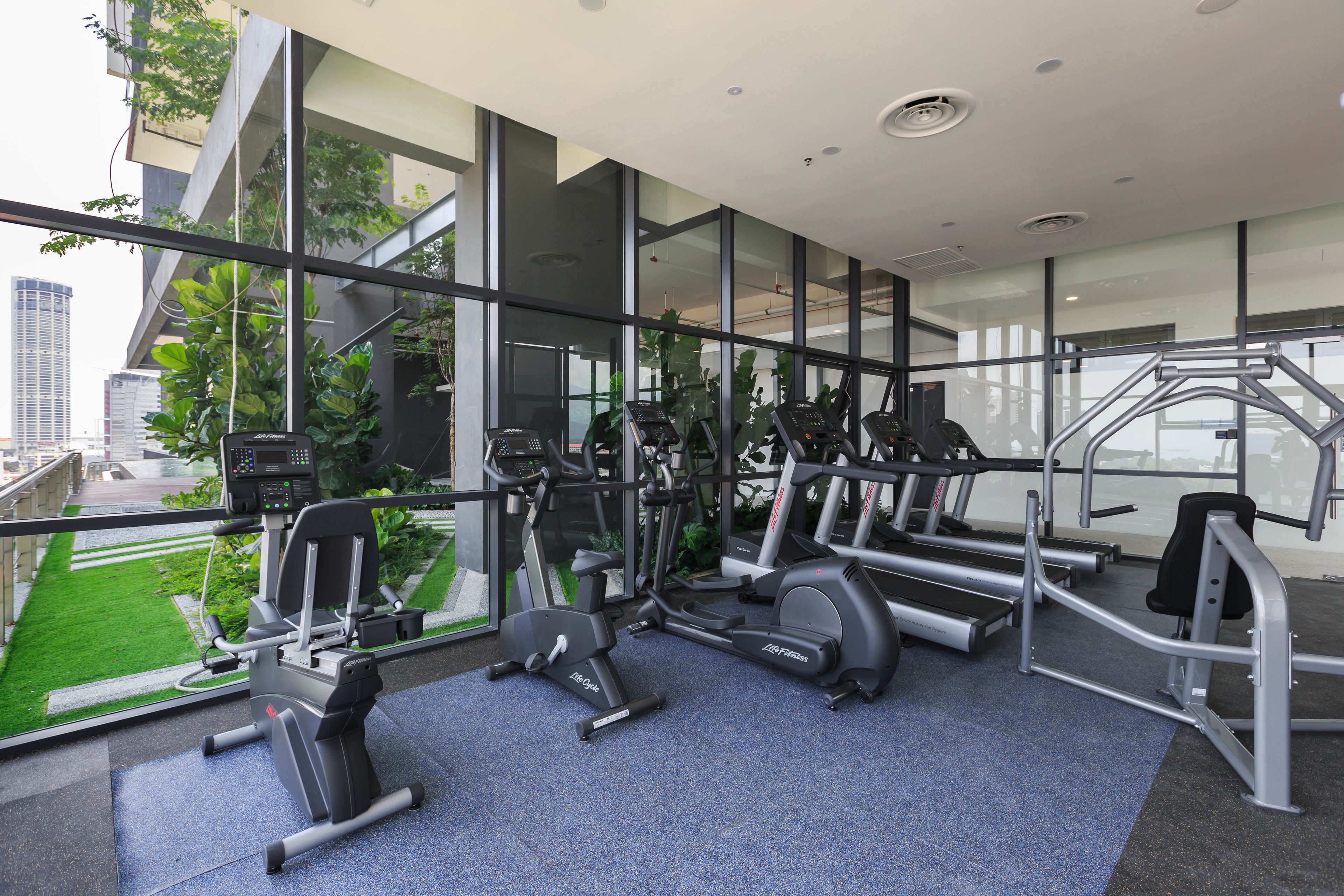 Un gimnasio moderno con diversos equipos de ejercicio, ventanas amplias y vistas a un paisaje urbano.