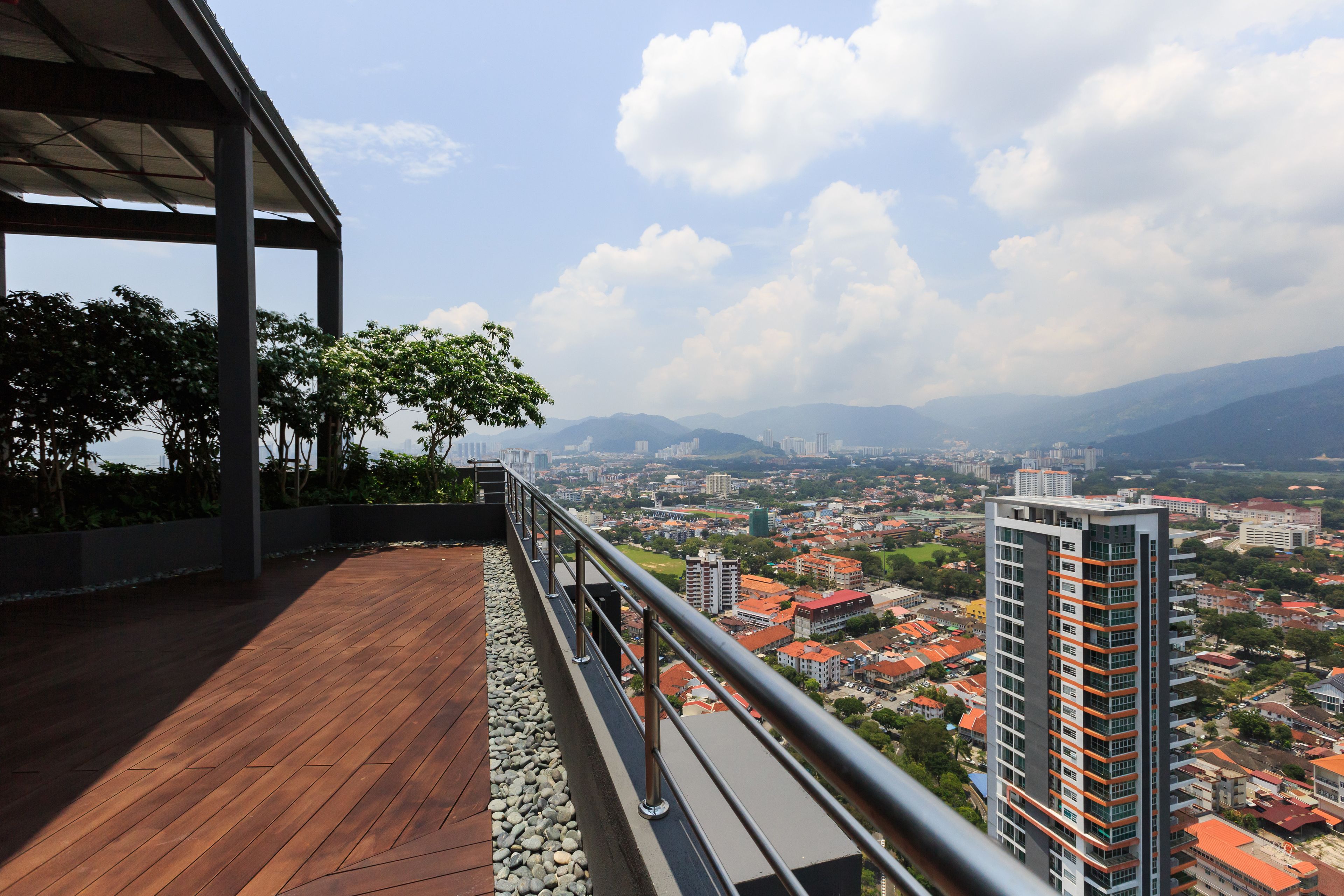 A balcony with a wooden floor and metal railing overlooking a cityscape with buildings and mountains.