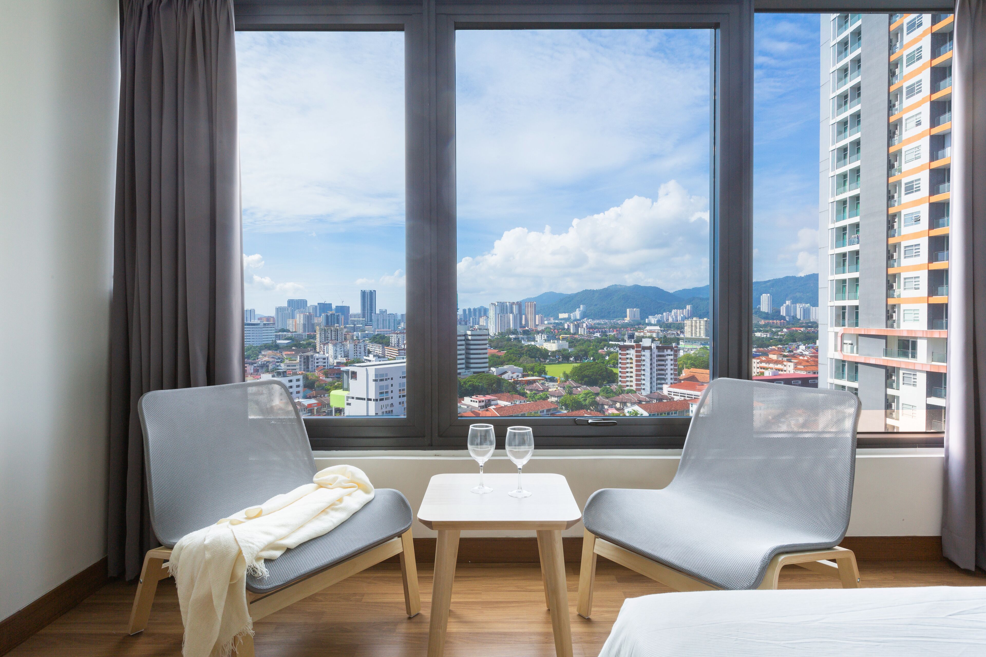 Hotelkamer met een groot raam met uitzicht op de stad, twee grijze stoelen, een kleine tafel met twee wijnglazen en een bed.