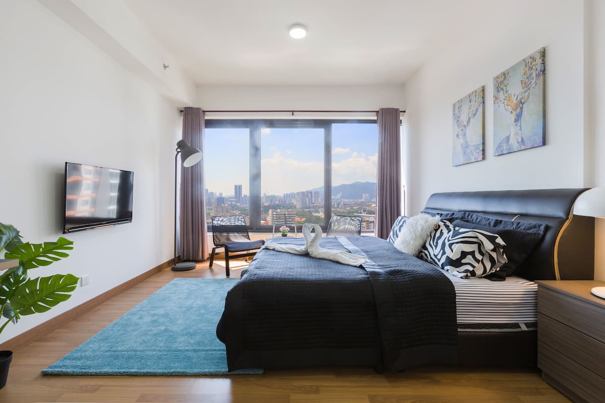 A modern bedroom with a large bed, a flat-screen TV, a city view, and a balcony.