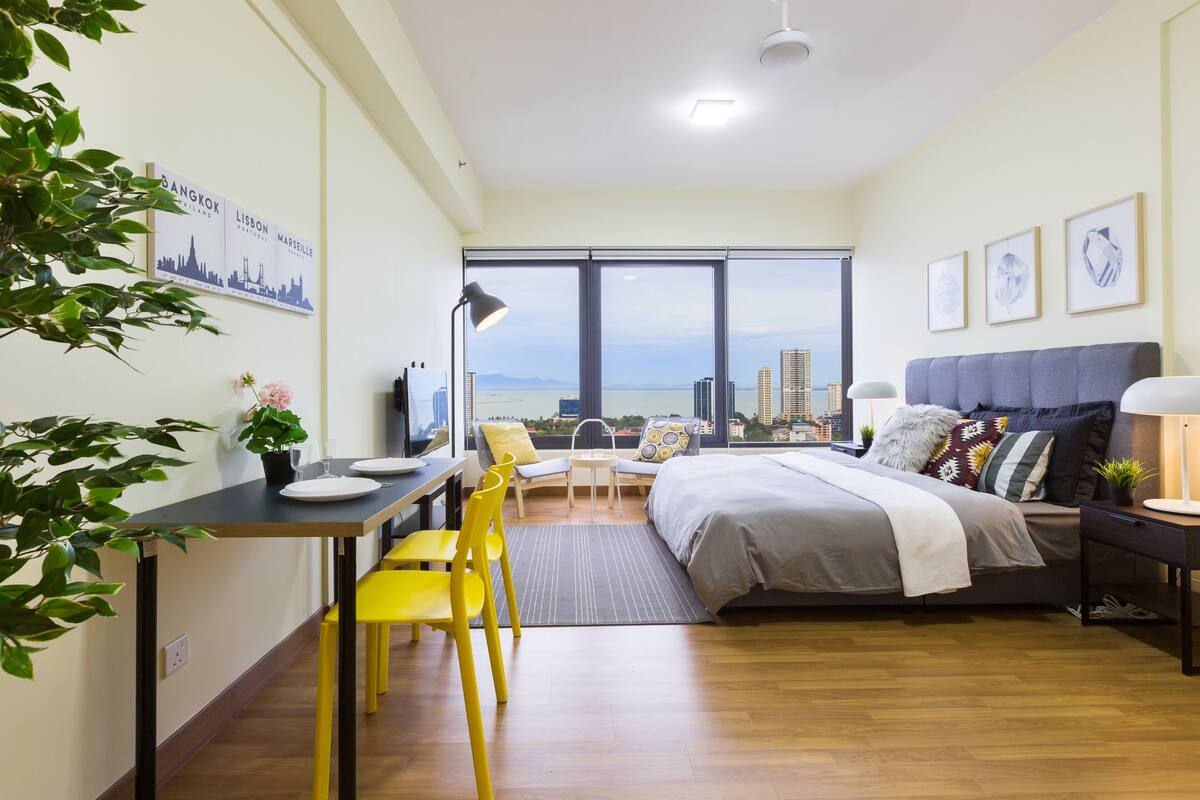 A modern bedroom with a city view, a dining area with yellow chairs, and a large bed with multiple pillows.