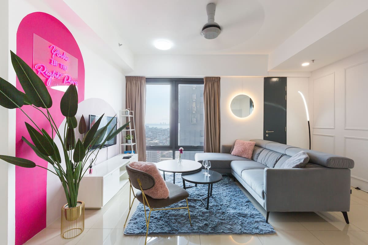 A modern living room with a grey sofa, a round coffee table, and a blue rug. There is a large window with curtains, a potted plant, and a neon sign on the wall.