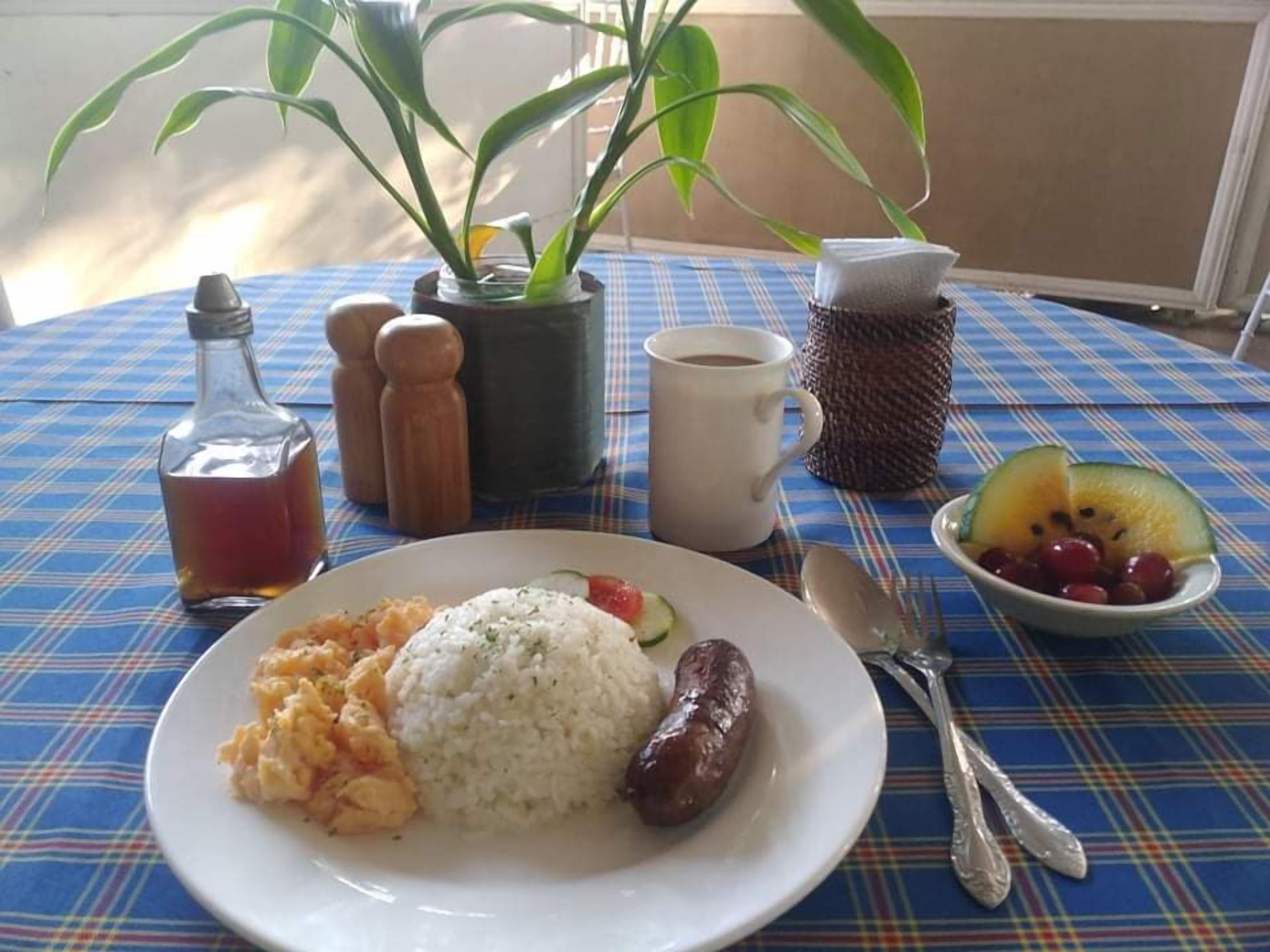 Uma mesa com toalha xadrez, um prato de comida, uma xícara de café, saleiro, pimenteiro, uma garrafa, uma tigela de frutas e uma planta em vaso.