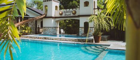 A swimming pool with a sign for Bella Vista Resort, surrounded by palm trees and a stone building with a tiled roof.
