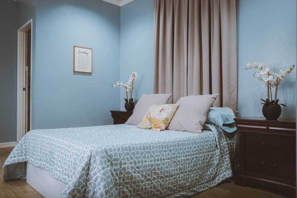 Une chambre à coucher comprenant un lit, deux tables de chevet, une commode et un mur orné d’une citation encadrée.