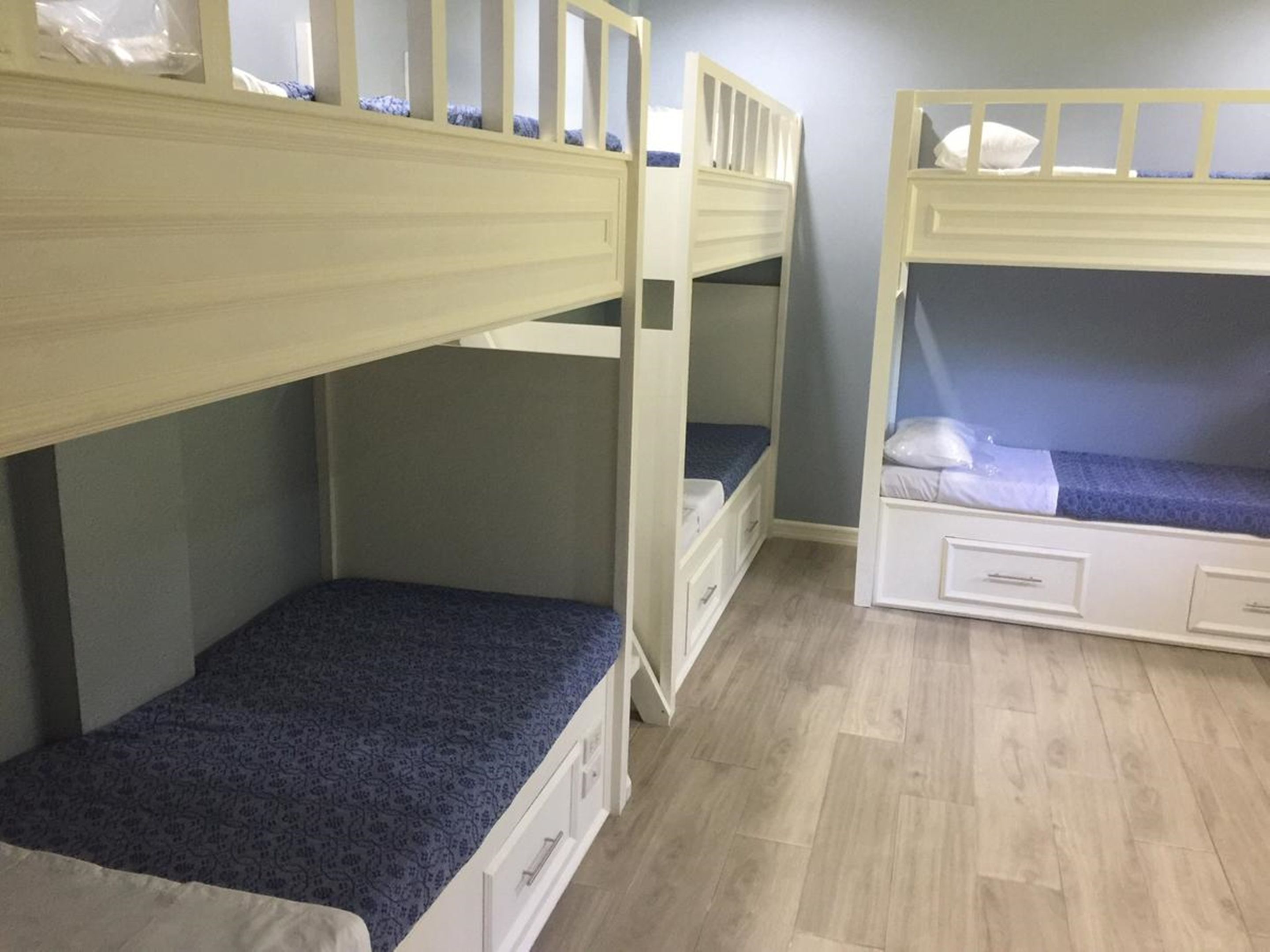 A room with three bunk beds, each with a mattress and a small drawer. The room has wooden flooring and light-colored walls.