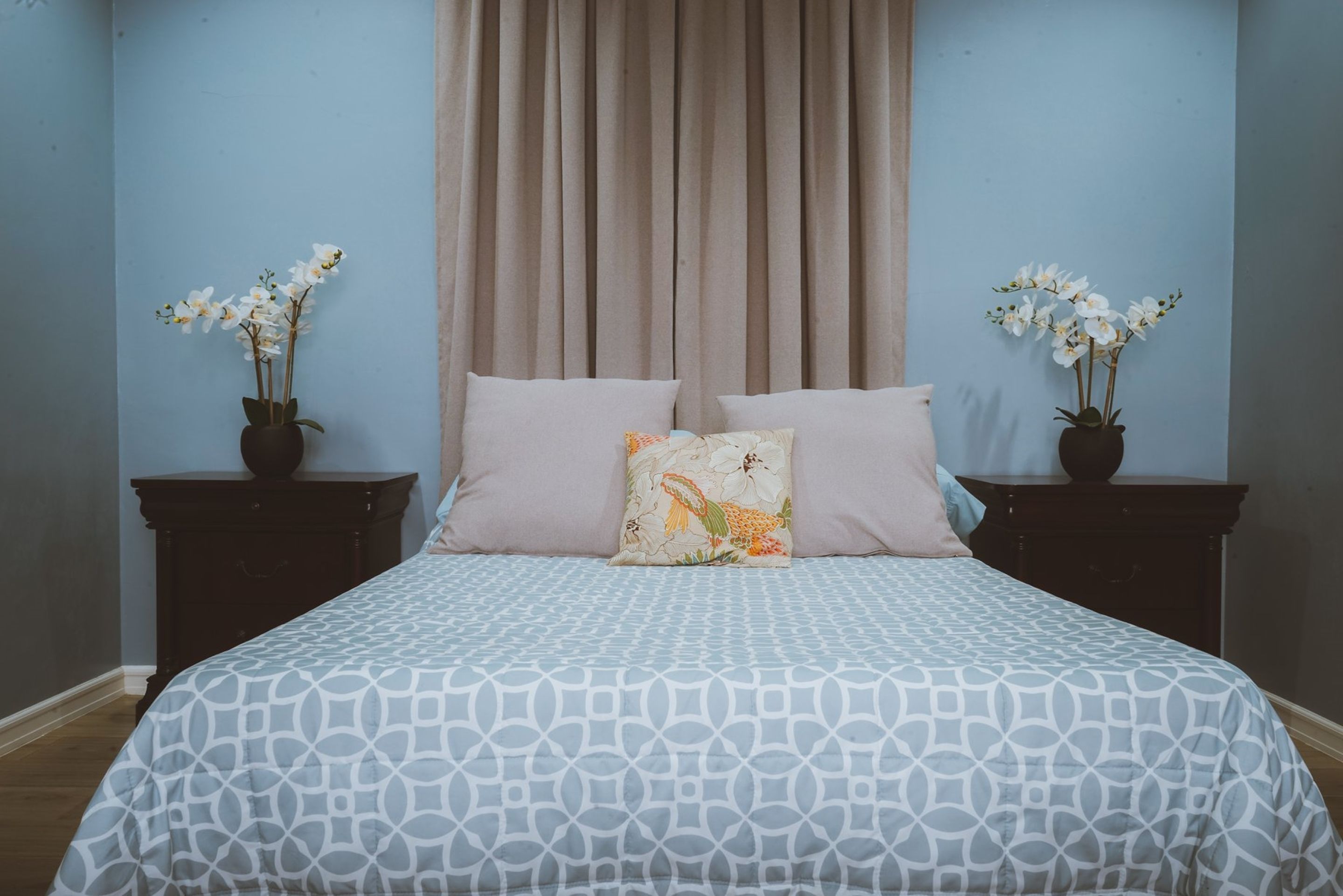 A bedroom with a patterned bedspread, two nightstands with potted orchids, a beige curtain, and a pillow with a fish design.