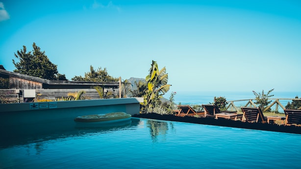 A pool with lounge chairs and a wooden deck, surrounded by tropical plants and a view of the ocean. - Chalets Chamarel (Chamarel)