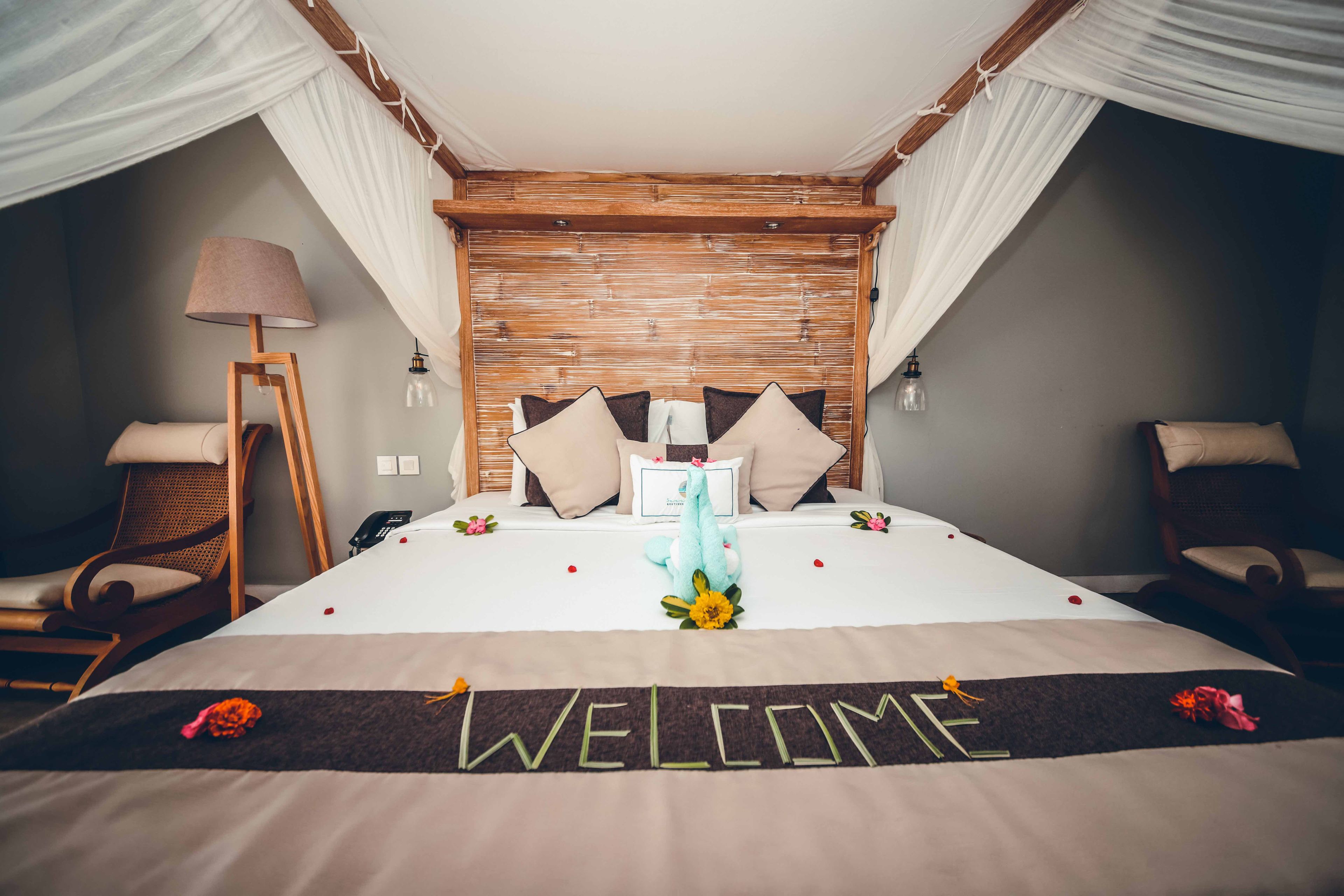 A bed with a 'WELCOME' sign, pillows, a lamp, and a chair.