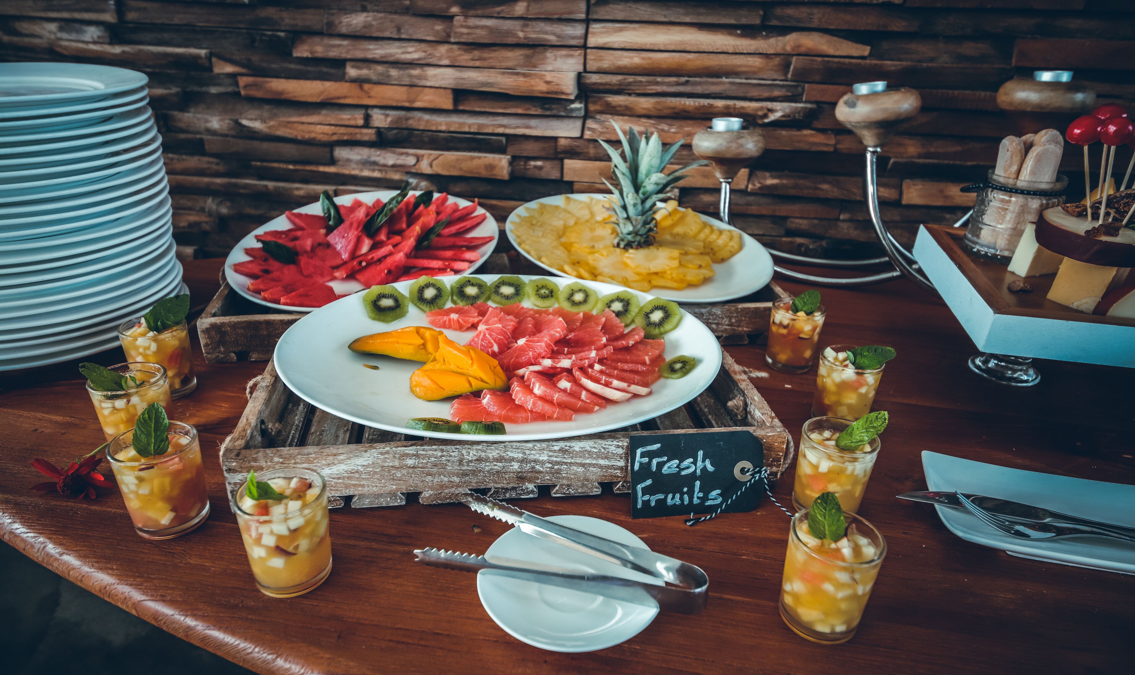 A buffet table with various fruits and drinks, a sign that says "Fresh Fruits", and a rustic wooden background.