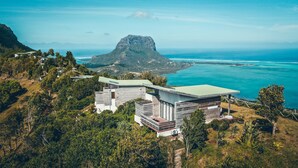 A modern house with a flat roof, surrounded by lush greenery and overlooking a coastal area with a prominent mountain in the background. - Chalets Chamarel (Chamarel)