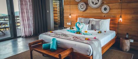 Habitación de hotel con cabecera de madera, cama con almohadones decorativos, un banco de madera y vista a través de un ventanal.