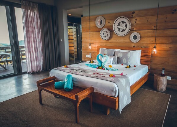 Habitación de hotel con cabecera de madera, cama con almohadones decorativos, un banco de madera y vista a través de un ventanal.