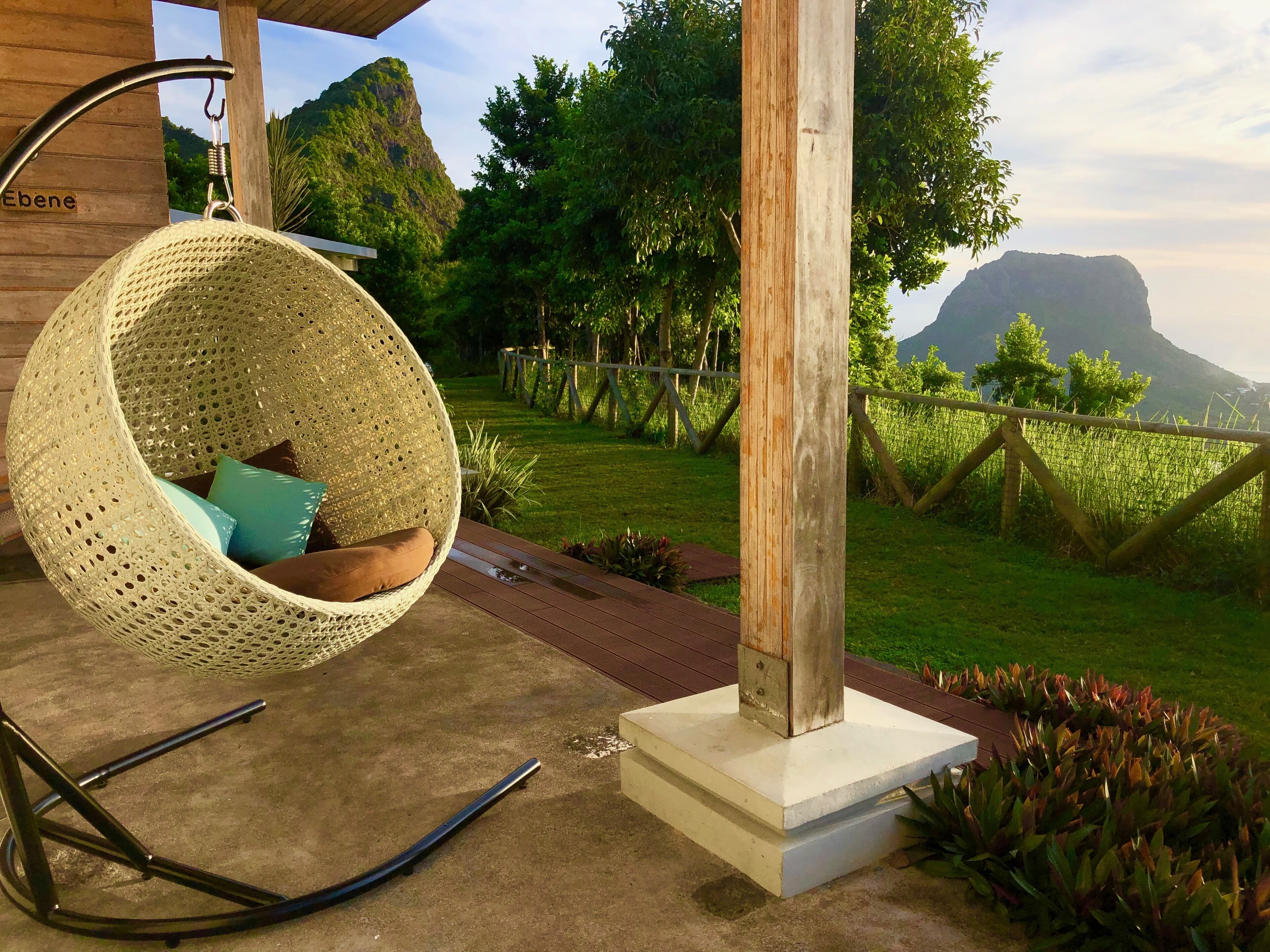 Un fauteuil suspendu avec des coussins, une terrasse en béton, un poteau en bois et une vue sur une zone verdoyante avec des arbres et une montagne en arrière-plan.