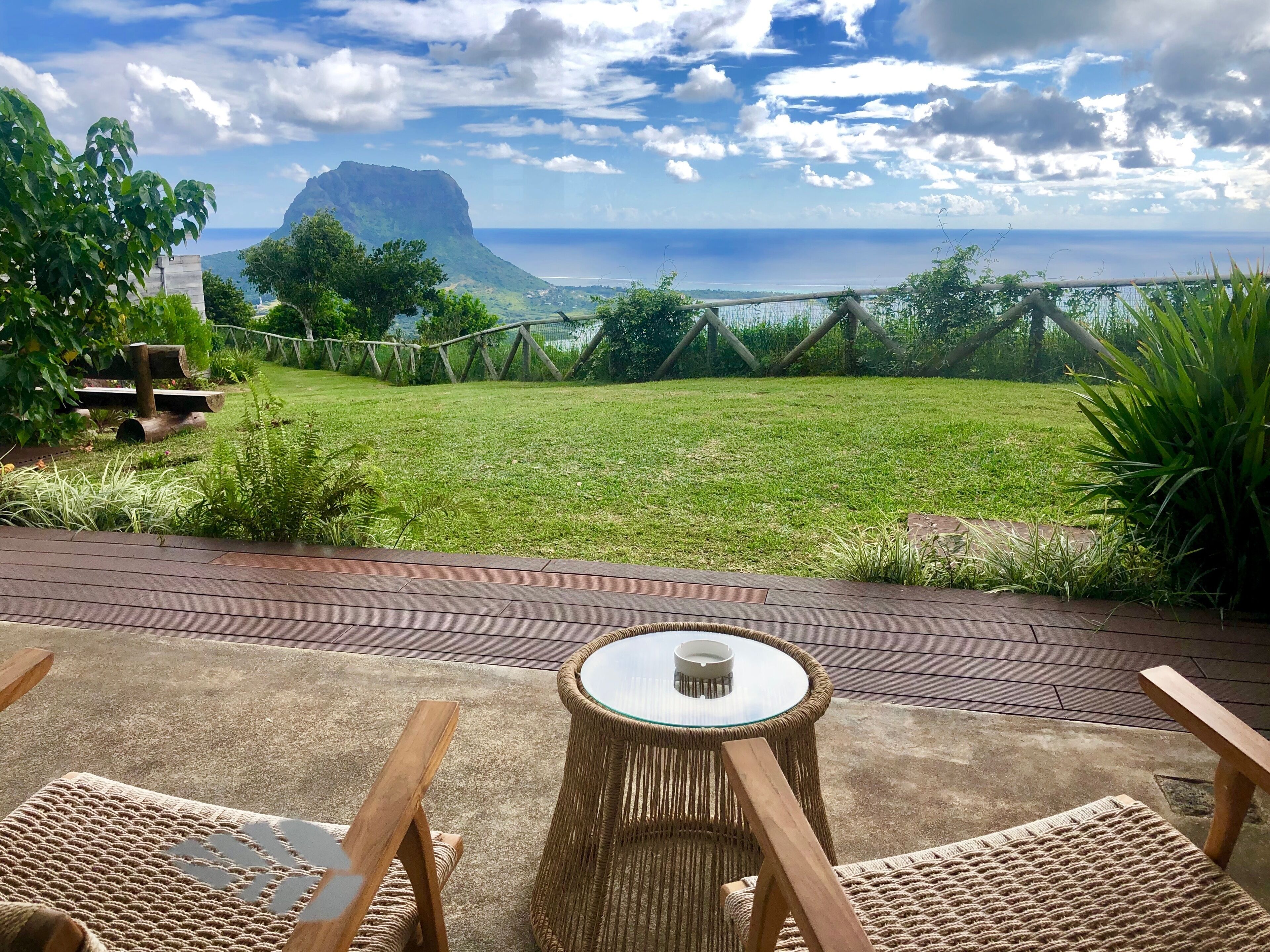 Une terrasse dotée de meubles en osier et d’une table en verre, offrant une vue sur une pelouse et l’océan.