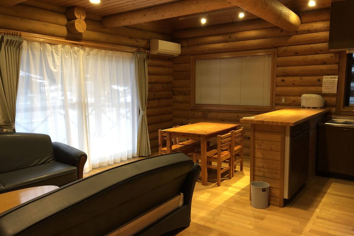 A log cabin interior with a sofa, dining table, chairs, a small kitchen area, and a window with curtains.