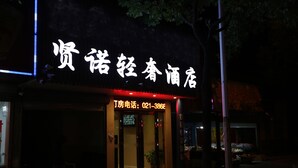 Ein Hotel Eingang mit einem leuchtenden Neonzeichen in einer fremden Sprache, einer gut sichtbaren Telefonnummer und einer kleinen Vorhalle mit einem Blumentopf.