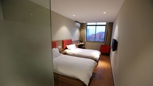 Ein Hotelzimmer mit zwei Betten, einem kleinen Schreibtisch, einem Stuhl und einem Fenster mit Blick auf die Stadt.
