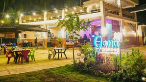 An outdoor dining area with tables and chairs, a neon sign, and a building with balconies. - Gana Siargao Island Resort (General Luna)