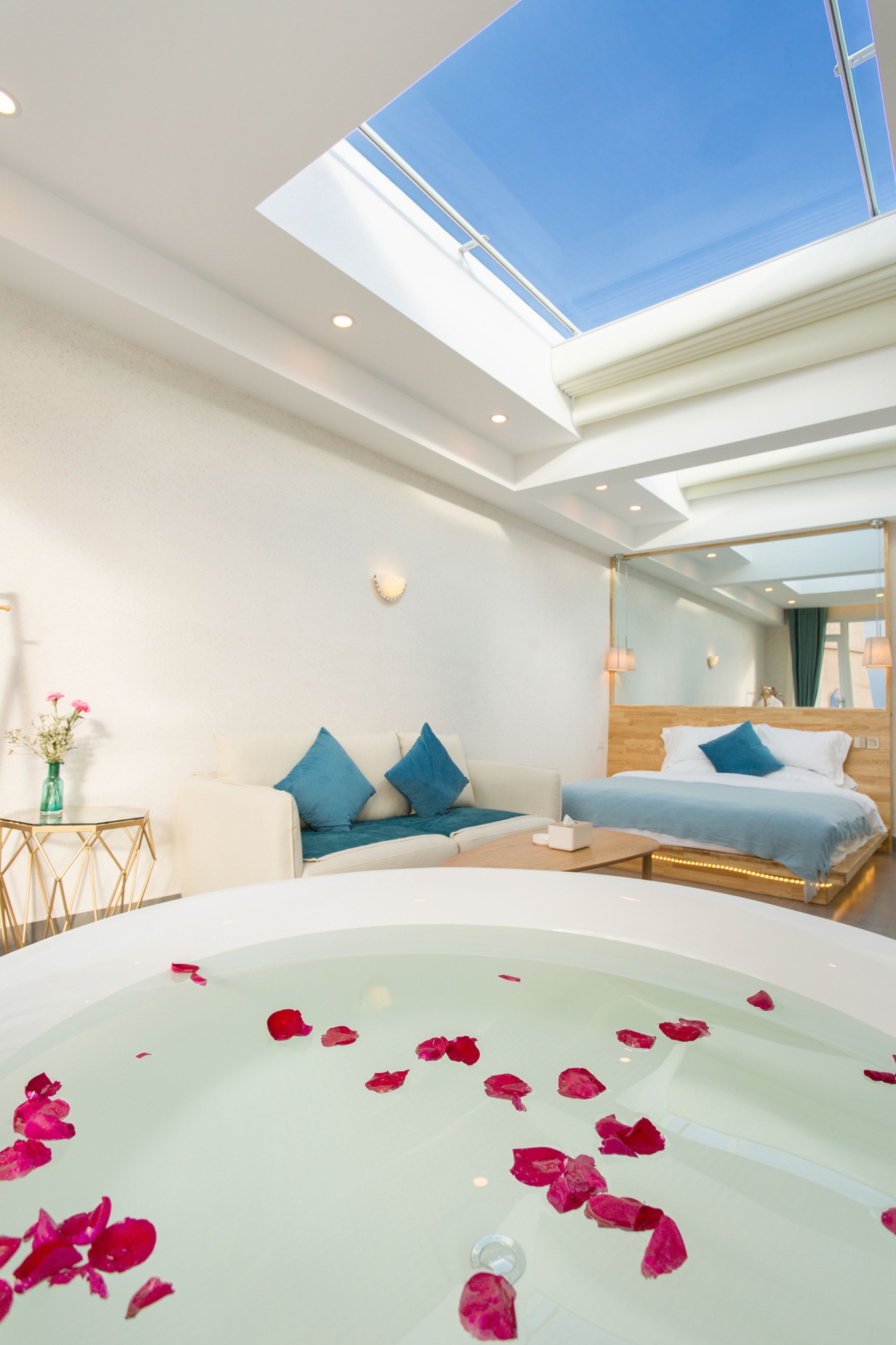 A modern hotel room with a skylight, a large bed with blue bedding, a sitting area with a white sofa, and a bathtub with rose petals.