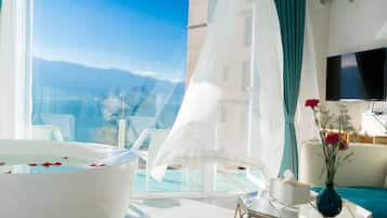 A modern hotel room with a glass-enclosed balcony, a white bathtub with rose petals, a flat-screen TV, and a vase with flowers.