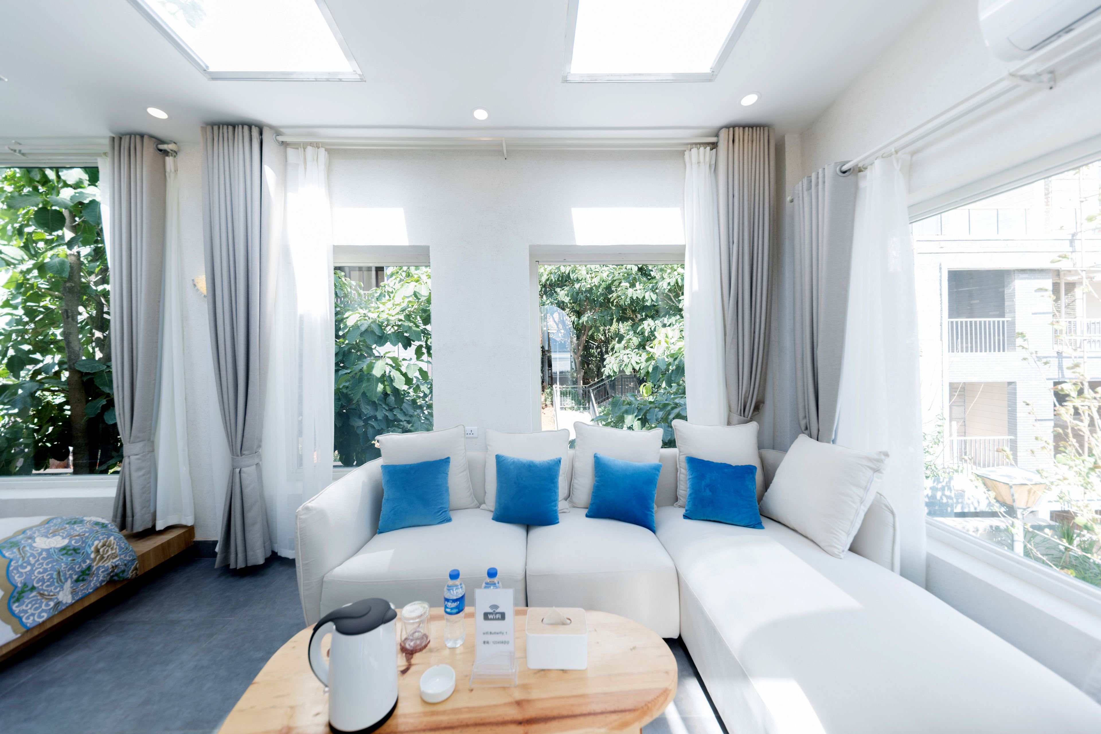 Habitación de hotel moderna con sofá blanco, cojines azules, una mesa de centro de madera y ventanas amplias con vistas a zonas verdes.