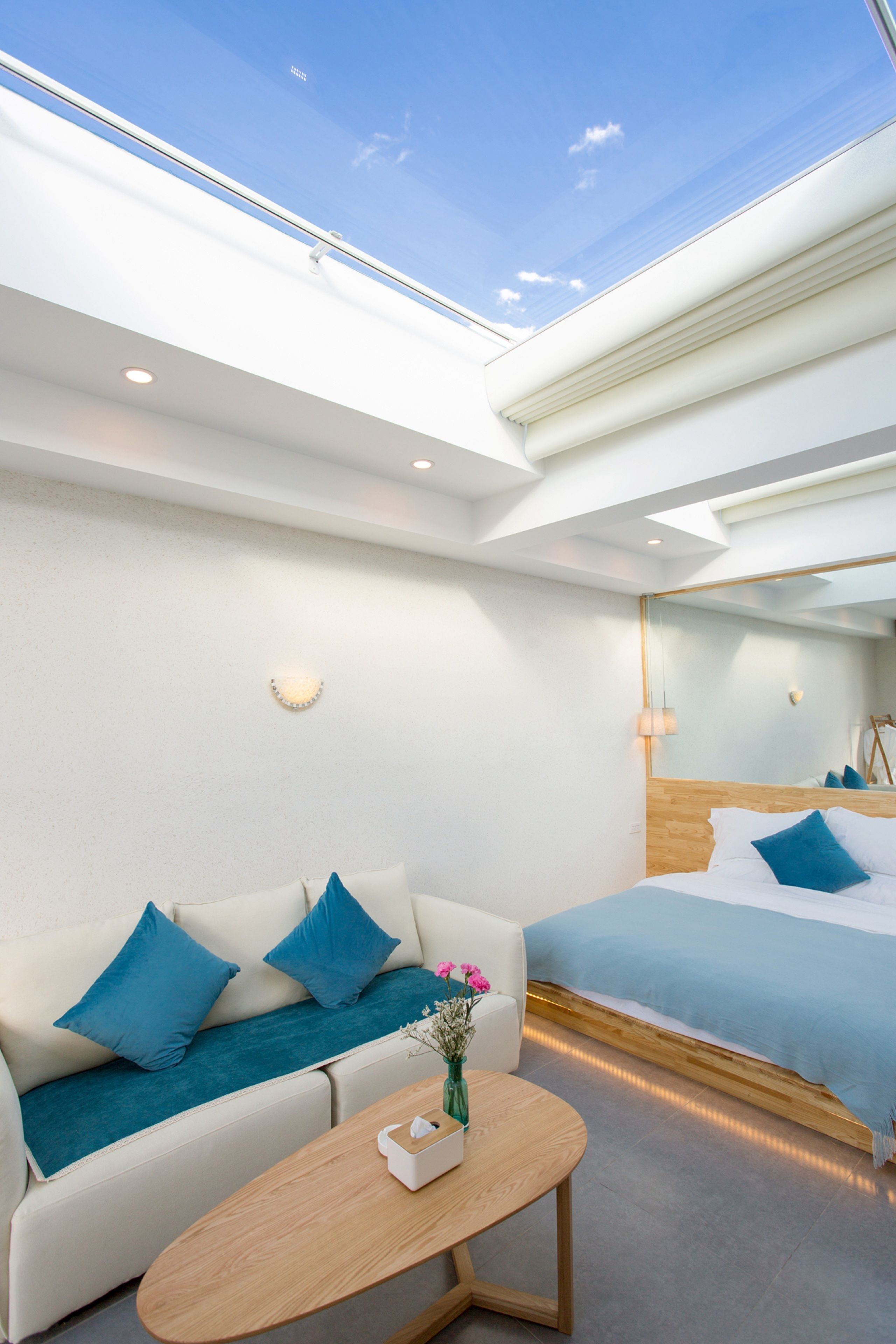 Une chambre d’hôtel moderne avec une lucarne, un lit avec une literie bleue, un canapé avec des coussins bleus et une petite table sur laquelle est posé un vase de fleurs.