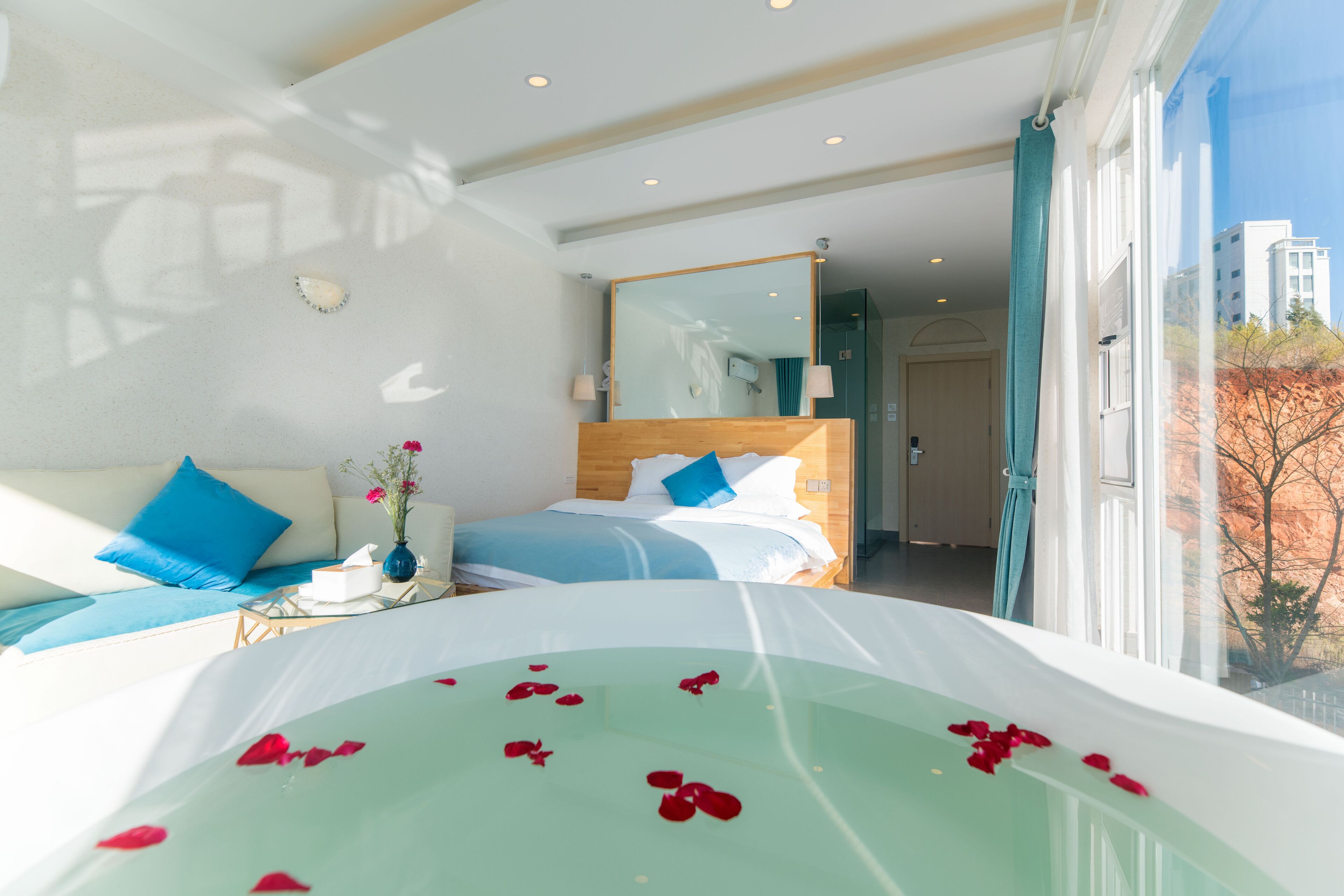 Habitación de hotel con una cama grande, un jacuzzi con cubierta de vidrio y pétalos de rosa, un cabecero de madera y vista a la ciudad.