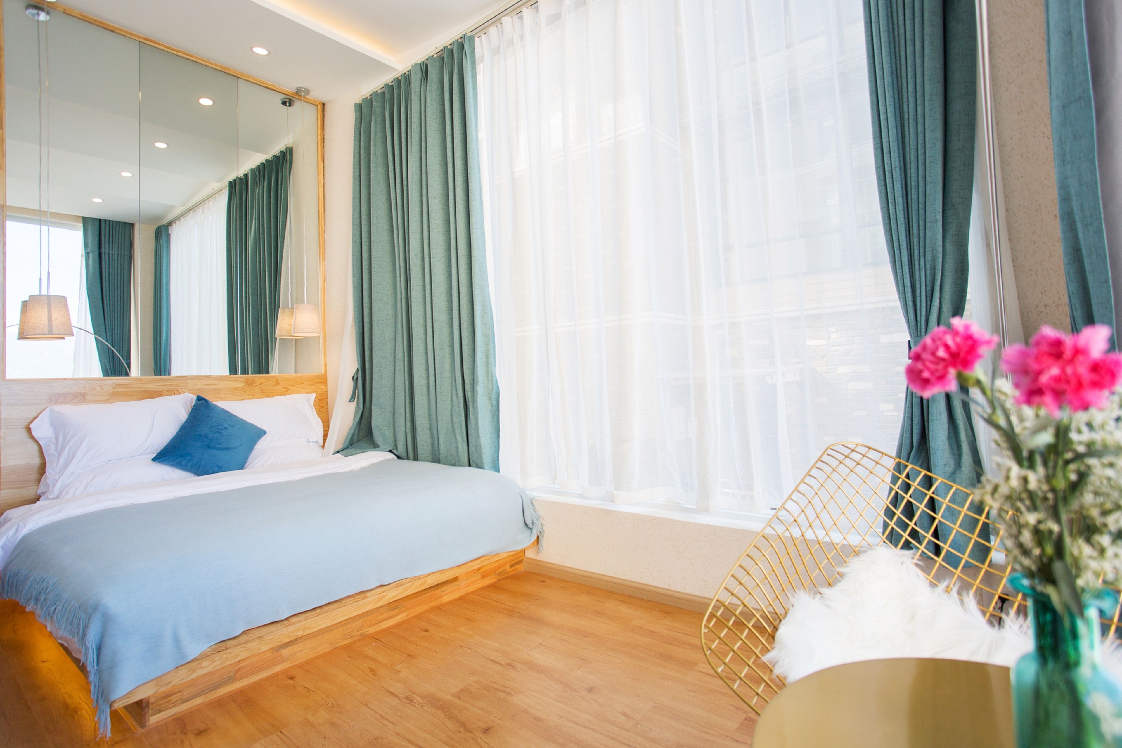 A bedroom with a bed, a mirror, a window with curtains, a chair, and a vase with flowers.