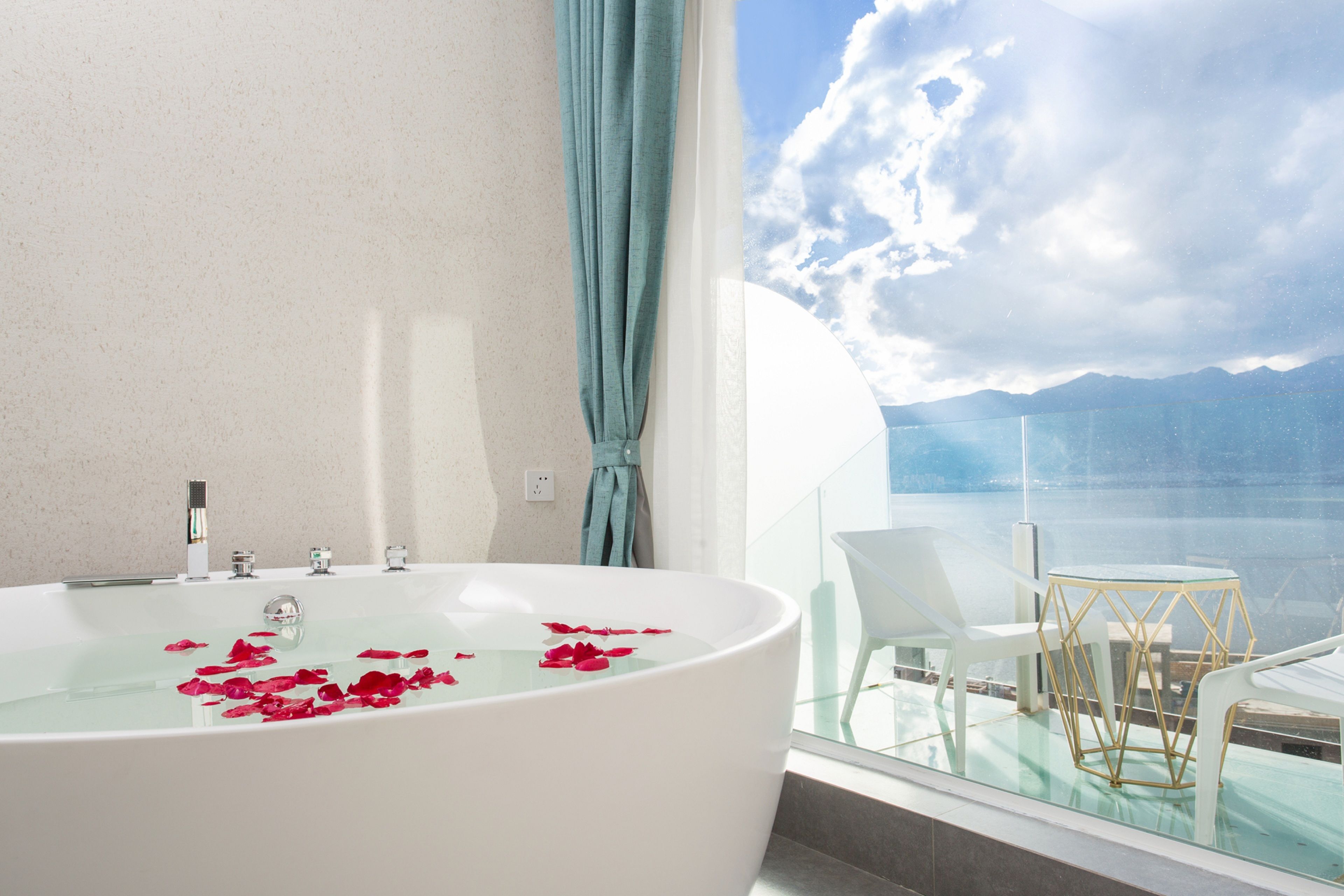 A modern bathroom with a large bathtub, a glass-enclosed balcony with a view of the sea, and a white chair.