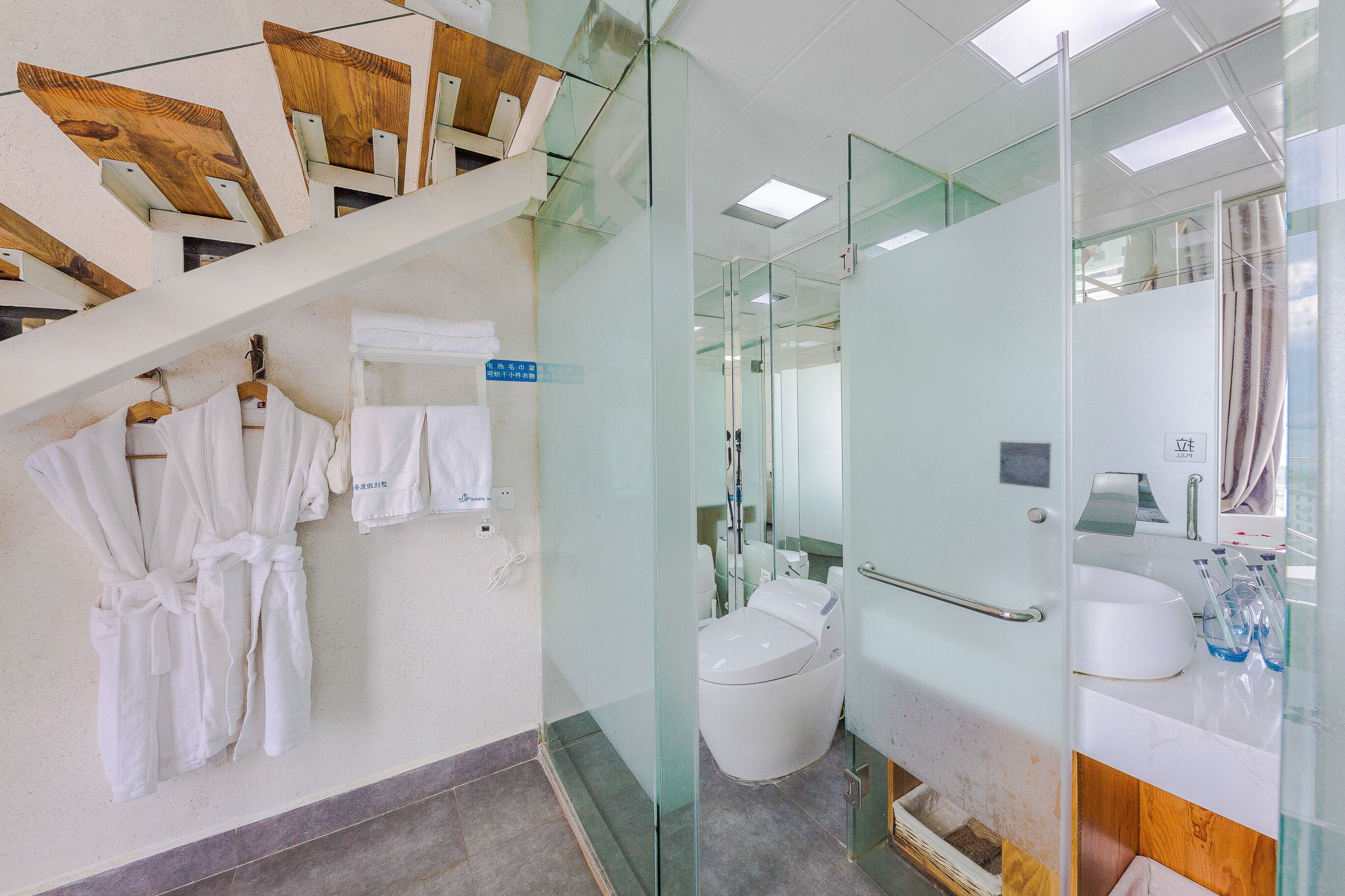 Uma casa de banho moderna com paredes de vidro, sanita branca e lavatório com artigos de higiene pessoal.