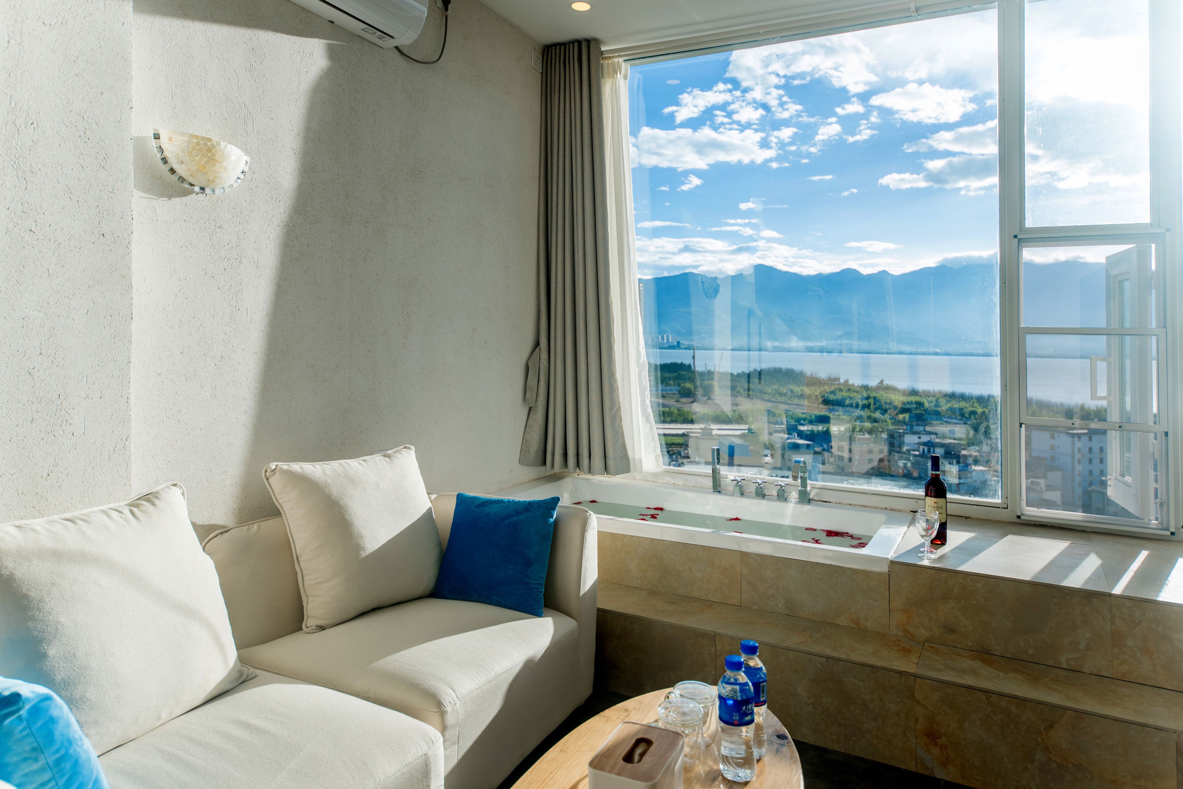 A hotel room with a large window, a sofa, a small table with bottles and glasses, and a view of a lake and mountains.
